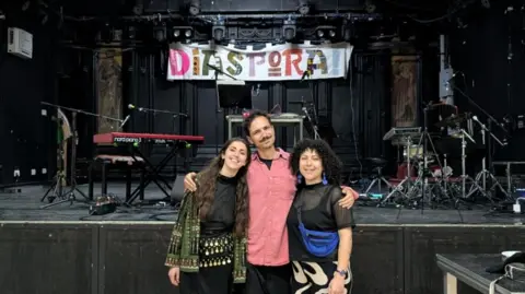 Makaani Arts A man and two women pose in from of an empty stage with the word 'Diaspora' hanging above. 