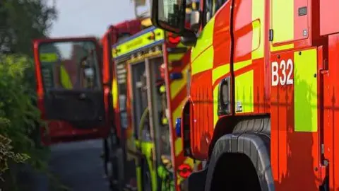 Stock image of the side of two fire engines. The one in the background has its door open to the left, towards a green, leafy hedge. The nearest engine is red and hi-visibility yellow with B32 in white font on its side. There is also the top of a black wheel arch. 