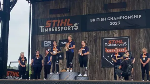 Justine Ellis Three women stand on podiums, holding trophies and wearing medals. There are other women stood behind them on a wooden stage.