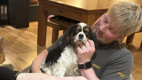 Matt Grimshaw Brown, white and black cavalier King Charles spaniel being held by her owner. They are sitting in a room with wooden floorboards and are sitting in a low chair. Matt is stroking the dog.