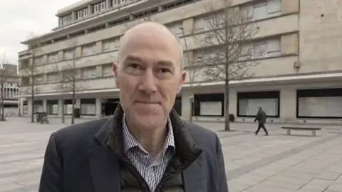 A man called Harry Holt stands outside a former department store in Plymouth city centre. The building is large and appears to be made from sandstone blocks. Holt is a bald man wearing a black suit jacket over a dark Barbour jacket and collared shirt.