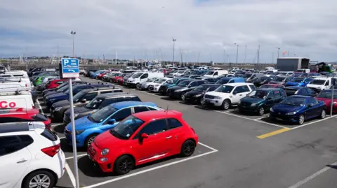 Lots of cars parked in a coastal car park in Guernsey. There are rows of cars packed in marked bays in a concrete car park. 