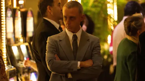 Studio Canal Jeremy Strong as Roy Cohn in The Apprentice - he is surrounded by slot machines and is wearing a gray suit and has his arms crossed