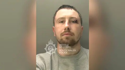 South Wales Police A man stood being photographed for a police mugshot. He has a gash above his left eye and is wearing a grey jumper.