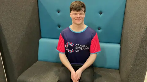 A person wearing a Cancer Research UK running shirt sits on a cushioned bench with a blue and grey backdrop.