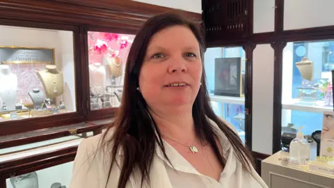 Lisa is looking into the camera and smiling slightly; she is wearing a white collared shirt and a white blazer. She is standing inside the jewellers, with glass cabinets behind her displaying necklaces and earrings. The room is painted white with dark wood panelling. 