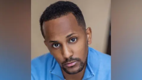 Dujonna Gift An actor's publicity shot. His face is inclined slightly to the right. He has a brown closely clipped beard and a buzz cut and is wearing a blue quarter-zip jumper.