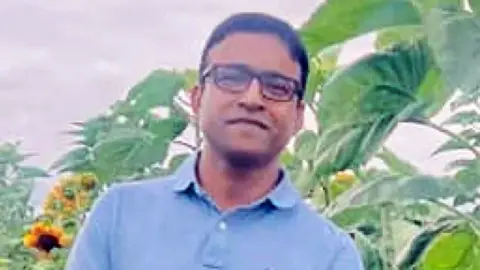 A man in his 40s with short, black hair wearing a blue polo shirt and glasses standing in front of plants