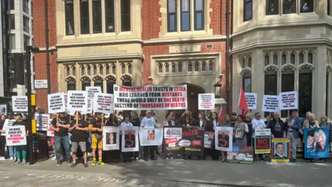 PA Media Around 30 families stand outside Arundel House in london protesting about the number of deaths in mental health services in Essex. They are holding banners, and pictures of their loved ones. Arundel House in London is a red brick mock tudor style building