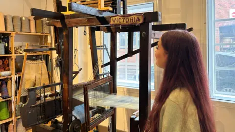 Charlotte stands next to a Victorian handloom and there are yarn supplies on the shelves behind it.