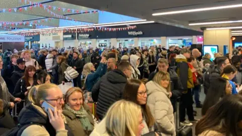 Grace Wood/BBC A crowd of people stood next to the ticket gates at Leeds Station as they are unable to get through the ticket barriers on to the platforms. Some are looking at their phones while others stand staring ahead. 