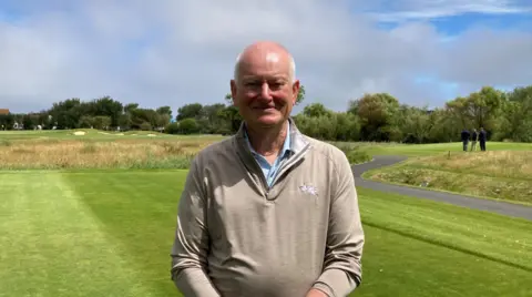 Stephen Lansdown, an older man, looking into the camera and smiling. He is wearing a biege quarter zip shirt and is standing in front of a golf course