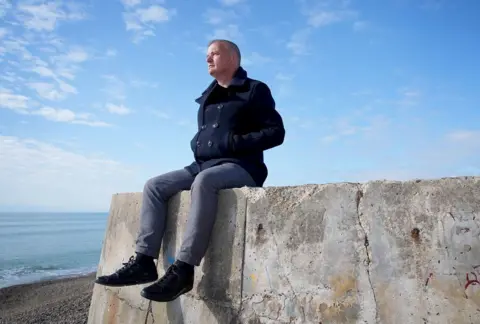 PA Craig Jones sat on a stone walls looking out to sea on a beach