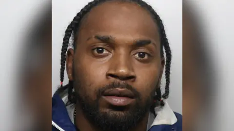 A police headshot of a Ranei Wilks. He has black dreads and is wearing a blue and white waterproof jacket. 