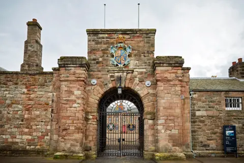 English Heritage Brick and stone gate house building with coat of arms, two chimneys and a double metal gate. There is a window with bars on it to the right of the entrance.