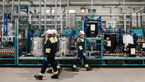 Three people walking inside of the Lithium Hydroxide Demonstration plant. There are two people walking to the left wearing white hard hats, black overalls with hi-vis around the neck, arms and feet. There is a person walking the opposite way wearing the same overalls. Behind the people are blue, silver and black metal structures with equipment on it. 