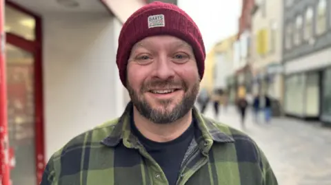 Ian Warborn has a dark beard and is smiling standing in a high street. He is wearing a red beanie hat and a green-and-black plaid shirt.