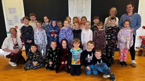 Bratton Primary School A group of children and staff at a primary school, wearing pyjamas and fluffy dressing gowns, standing in the school hall and posing for a photo.