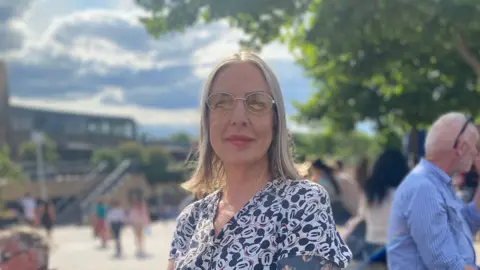 Dr Marsha Smith Dr Marsha Smith, a White woman with blond hair wearing glasses that have slightly yellow-tinted lenses. She is wearing a printed blouse covered in Mickey Mouse faces and has left arm is covered in tattoos.