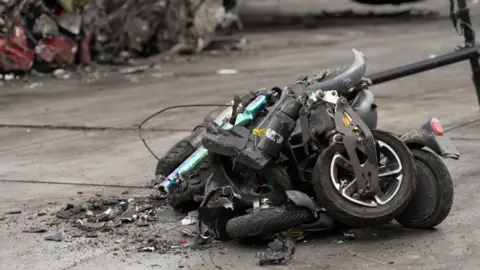 BBC The image shows a pile of crumpled up e-scooters and e-bikes. They are all black in colour apart from one which is a two toned green and purple colour. 