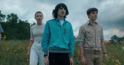 Three young people, two males and a female with a shaved head, stand in a field with long grass and various colours. They're looking out at something off-camera in front of them, with expressions of concern and worry.