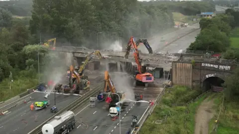 BBC A number of bulldozers start to bring down the bridge