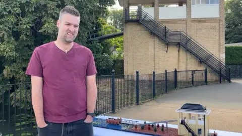 Wicksteed Park A man in a purple t-shirt and jeans stands next to a Lego model of a water ride. He is standing next to the real life version of the attraction he has built. 