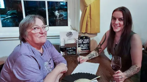 Lynda (left) with short grey hair wearing a lilac t-shirt and glasses with her daughter Lydia with long dark hair wearing a black sleeveless top sitting at a table in a pub by the window with a drink and pen in hand.