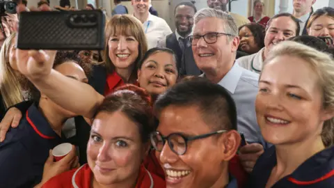 Close up Sir Kier Starmer and Rachel Reeves posing for a selfie taken by an NHS staff member. They are smiling and surrounded by a group of NHS staff all smiling at the camera