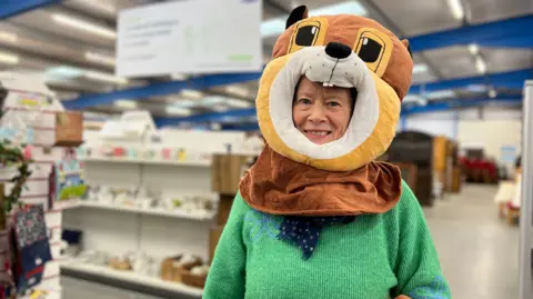 Caroline smiles at the camera while wearing a squirrel costume head. She has a green jumper on as well as a dark blue scarf. She's standing in the lane of a warehouse which is used to sell second hand items for the charity.