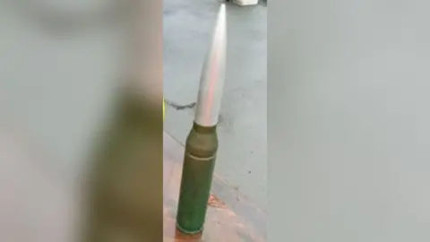 A military shell about 45cm tall sits on an outdoor surface. Its propellant section is brass coloured, with a large silver projectile on top. 