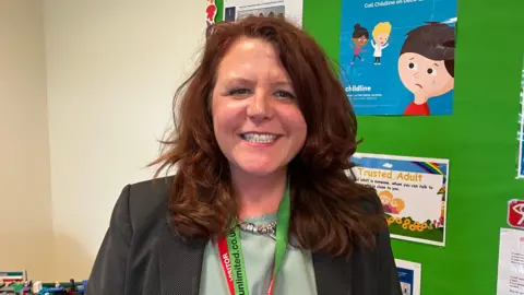 Michelle Palmer smiling at the camera with a wide friendly smile. She has red hair and is standing in front of a display board with a cartoon of a boy crying.