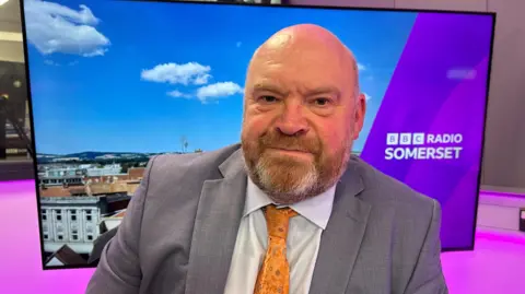 A man in a grey suit and yellow tie sitting in front of a large screen with purple BBC Radio Somerset branding shown