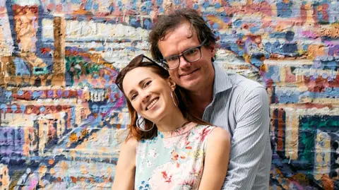 Francesca Baker-Brooker Photo of a couple with the man stood behind, hugging his partner. She wears a sleeveless floral top and sunglasses on her head, large gold hoop earrings, dark brown hair and he has dark brown hair, wears a blue and white striped top and spectacles. They both smile at the camera and are stood in front of a brightly coloured graffitied brick wall.