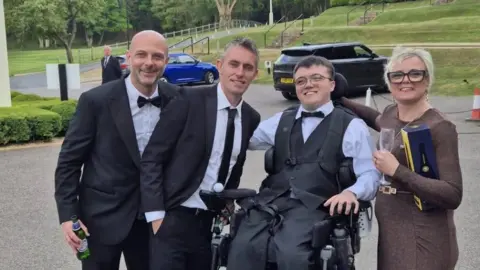 Harrison Mayhew-Kemp Three men and a woman stand together in a line to take a photo. One of the men is in a wheelchair. The men wear black suits, white shirts and ties while the woman is wearing a brown dress. They are all smiling.