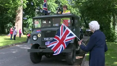 A vintage military vehicle passes an older woman waving a Union Jack flag at the event in 2019. Other vehicles can be seen behind it.