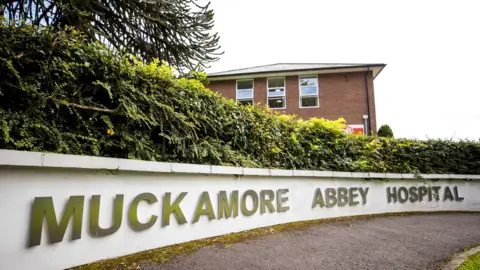 PA Media Muckamore Abbey Hospital - the name is in big capital letters along a low curved white wall. There is a path and patch of grass in front. There is a hedgerow behind it and then behind that a red brick building with three of the top windows visible behind.