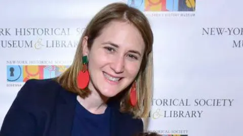 Getty Images Wesley LePatner poses at New York Historical Society Museum event. She is smiling and wearing a dark blue shirt and blazer and and large dangling red and green earrings. In the background is a backdrop for the museum showing its logo. 