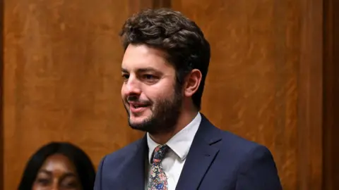 UK Parliament MP Jack Abbott speaking in the House of Commons. He is wearing a navy suit jacket, white shirt and has a patterned tie. He has dark hair and a beard.