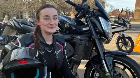 A woman with long brown hair, dressed in black motorcycle leathers with pink trim, is crouched in front of a black motorcycle. She is holding a black helmet.