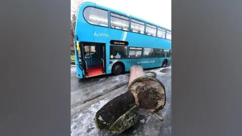 BBC A blue arriva bus next to a pillar which is has fallen down. There is ice and snow on the road and pavement.