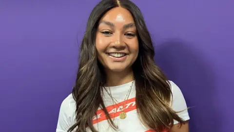 Lillian Istance pictured in front of a purple background. She has long brown hair and is wearing a white and red t-shirt with a gold necklace.