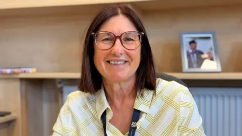 A women with short brown hair, glasses and a yellow and white patterned shirt. She is looking at the camera smiling.