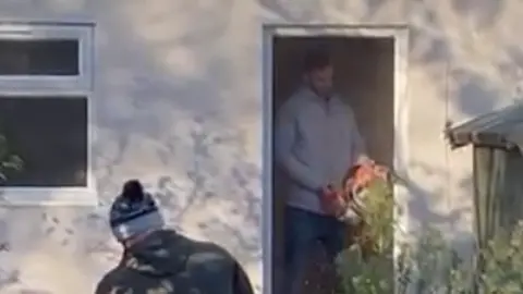 Gemma Hemmings A blurry image of Steve Figg holding an orange chainsaw. He is using it to damage the doorframe of a white garden office.
