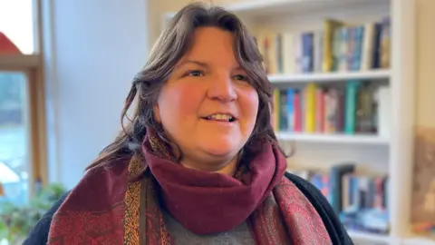 A woman with long, brown hair in a purple patterned scarf with a bookshelf in the background