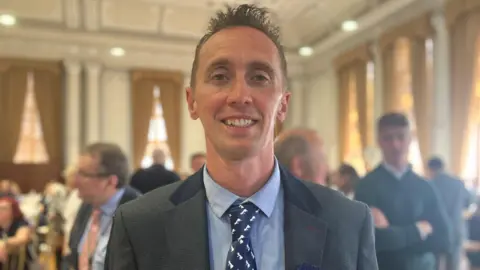 Owen Sennitt/BBC Jonathan Wedon a man who is standing having this picture taken and is looking directly at the camera and smiling. He is wearing a pale shirt with a patterned tie and blazer.