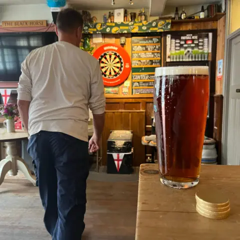 George Carden/BBC A man in a white top aims brass coins at a lead topped small table with a hole in it. In the foreground is a pint of beer and four brass coins