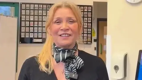 Cathy Hunt, a middle-aged woman with mid-length blonde hair, wearing a black neckerchief and a gold necklace with a cross pendant. She has a black blouse and stands in front of a grid showing small head and shoulders images of people.