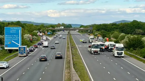 BBC/Andy Bennett Two sides of the motorway. You can see the Junction 22 sign and bridge above the lanes. On the right hand side the road is empty. There are several lorries and trucks surrounding the tanker which spilt fuel.