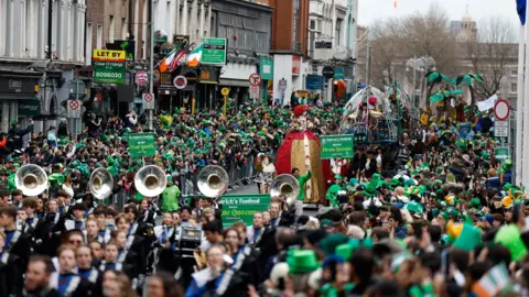 Reuters Crowds gathered in Dublin for St Patrick's Day parade. Floats are making their way down with street with bands playing large brass instruments.
